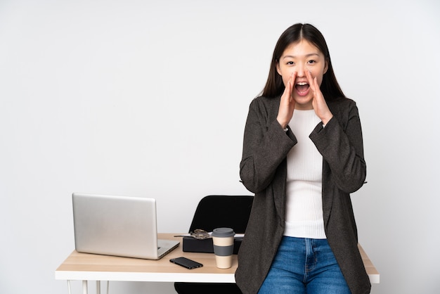 Mulher asiática de negócios em seu local de trabalho na parede branca, gritando e anunciando algo