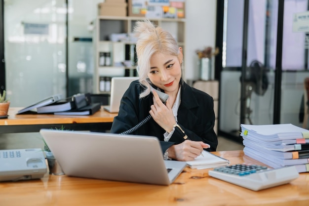 Mulher asiática de negócios falando ao telefone e usando um laptop com um sorriso enquanto está sentado no escritório