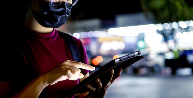 Mulher asiática de óculos usando uma máscara facial usando um tablet à noite na cidade