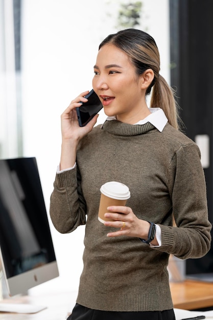 Mulher asiática de suéter segurando a xícara de café e usando smartphone em pé no escritório