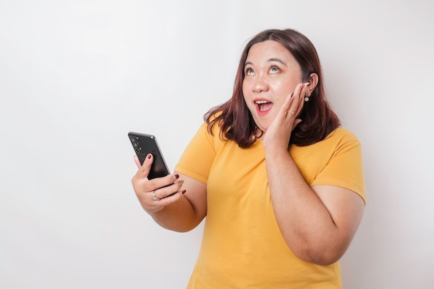 Mulher asiática de tamanho grande surpresa vestindo camiseta amarela apontando para seu smartphone isolado por fundo branco