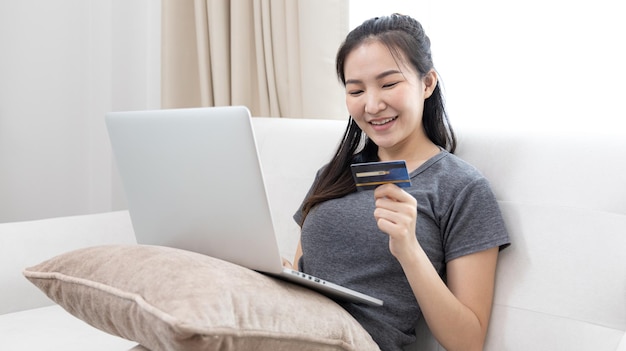 Mulher asiática descansando no sofá e usando laptop para fazer compras on-line com cartão de crédito para se registrar