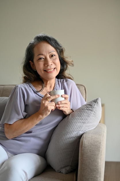 Mulher asiática dos anos 60 relaxando em sua sala de estar tomando seu café da manhã no sofá