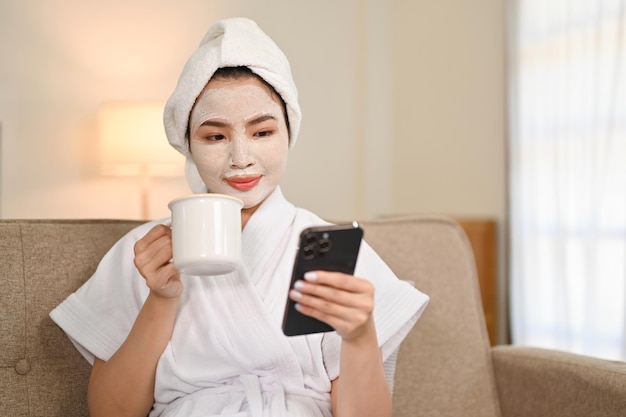 Mulher asiática em roupão e máscara facial de argila no rosto tomando café e usando o telefone