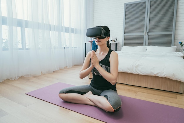 mulher asiática em roupas esportivas está usando óculos de realidade virtual enquanto medita com as palmas das mãos juntas no colchonete em uma manhã tranquila em casa com a luz do dia.