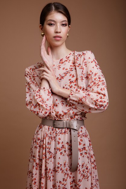 Mulher asiática em um vestido bege com padrão floral posando sobre um fundo de café marrom.