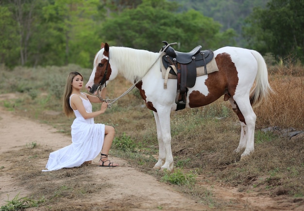 Mulher asiática em uma fantasia de vestido longo fica com um cavalo em uma fazenda de gado.