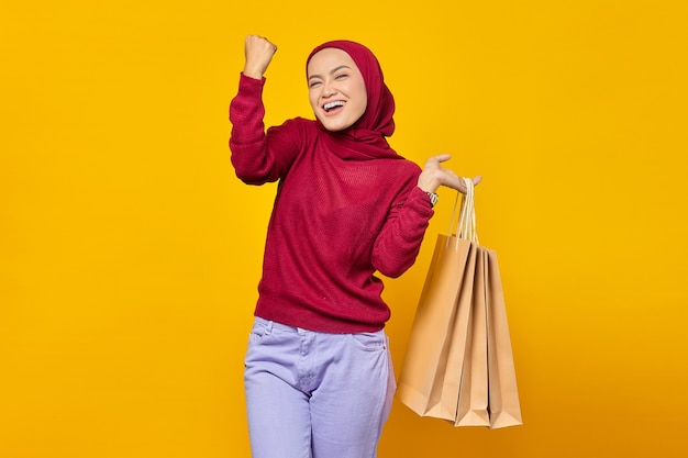 Mulher asiática empolgada comemorando o sucesso com uma expressão sorridente e mostrando sacolas de compras sobre fundo amarelo