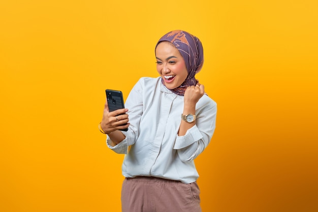 Mulher asiática empolgada olhando para smartphone recebendo boas notícias sobre fundo amarelo