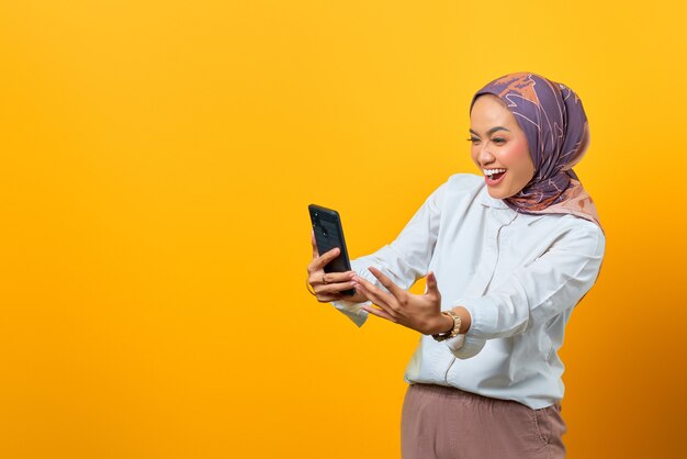 Mulher asiática empolgada olhando para smartphone recebendo boas notícias sobre fundo amarelo