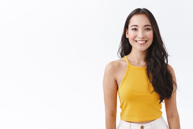 Foto mulher asiática entusiasmada da cintura para cima com longos cabelos escuros, usar um top amarelo da moda, sorrindo com uma expressão amigável e despreocupada como a câmera olhando, expressar emoções positivas e alegres, fundo branco