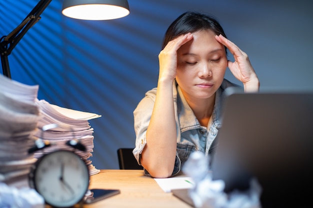Mulher asiática escrevendo documentos em horas extras à noite. prazo de trabalho no escritório. dor de cabeça de garota do oficial e ocupada com o trabalho. infeliz e estresse.