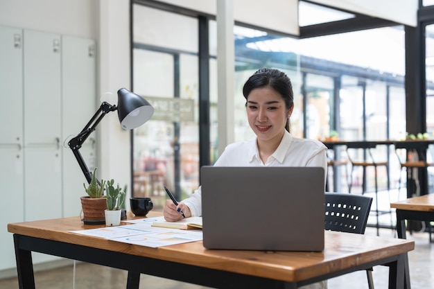 Mulher asiática escrevendo e anotando no notebook com laptop no escritório