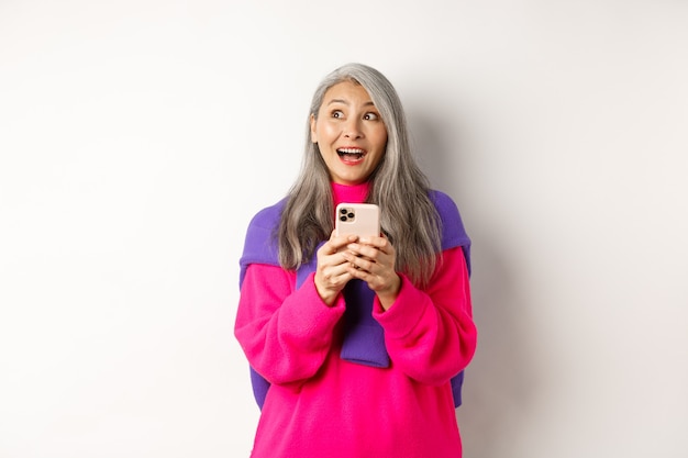 Mulher asiática espantada, sorrindo e olhando de lado fascinada depois de ler a promoção no smartphone, em pé com o telefone móvel sobre fundo branco.