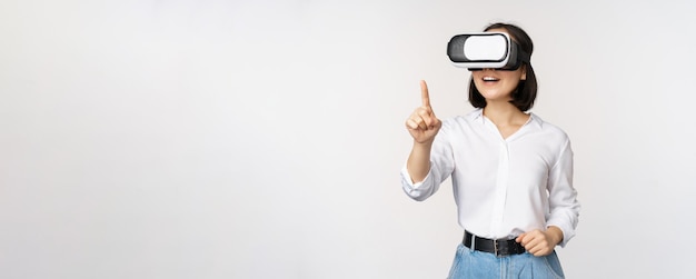 Mulher asiática espantada usando fone de ouvido vr garota coreana em óculos de realidade virtual apontando para smth e sorrindo fundo branco