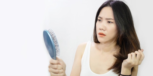 Mulher asiática está olhando para a perda de cabelo e segurando pente azul e cabelo com cara de preocupação e chocada