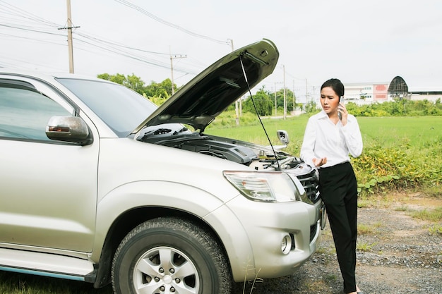 Mulher asiática estressada fica na frente do carro quebrado em seu trajeto para o trabalho, usando smartphone para obter ajuda.