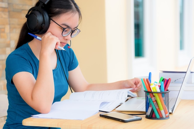 Mulher asiática estudante adolescente com óculos fones de ouvido sentado olhando sério lendo um livro se preocupe usando o computador portátil na mesa de aprendizagem de estudo on-line. educação de uma classe de universidade em casa