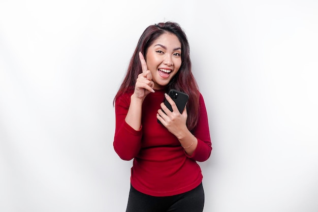 Mulher asiática excitada vestindo blusa vermelha apontando para o espaço da cópia em cima dela enquanto segura o telefone isolado pelo fundo branco