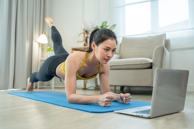 Mulher asiática exercitando ioga e assistindo tutorial online no laptop na sala de estar em casa