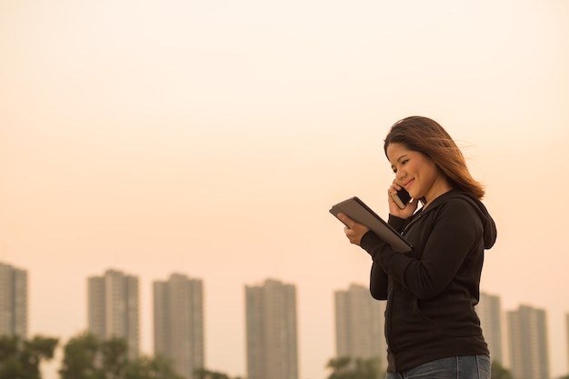 Mulher asiática falando no celular e segurando um tablet com o fundo do céu ao pôr do sol
