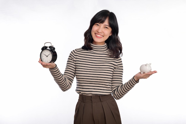 Mulher asiática feliz com camisa de suéter segurando despertador e cofrinho isolado no fundo branco