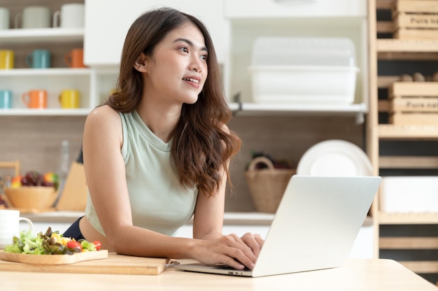 Mulher asiática feliz comendo salada com notebook de computador na sala da cozinha