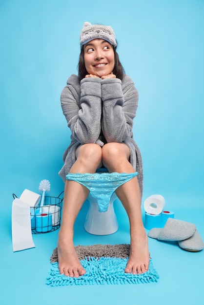 Mulher asiática feliz e sonhadora usando uma venda cinza e calcinha de renda de roupão nas pernas sente alívio enquanto está sentada no vaso sanitário posa contra a parede azul do banheiro