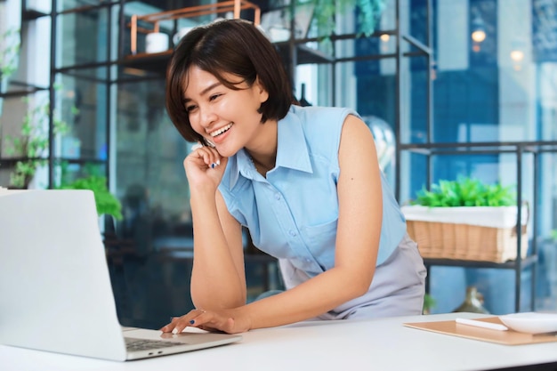 Mulher asiática feliz na camisa azul trabalhando com laptop no escritório