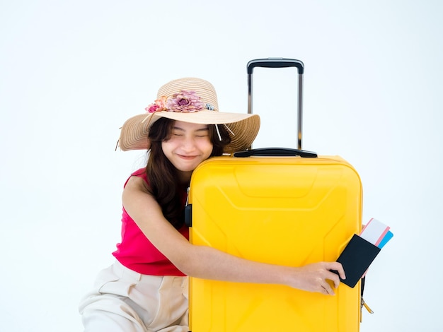 Mulher asiática feliz na camisa sem mangas rosa e chapéu de praia segurando a passagem de avião e abraçar a mala amarela isolada no fundo branco Jovem viajante pronto com animado para viagem de verão