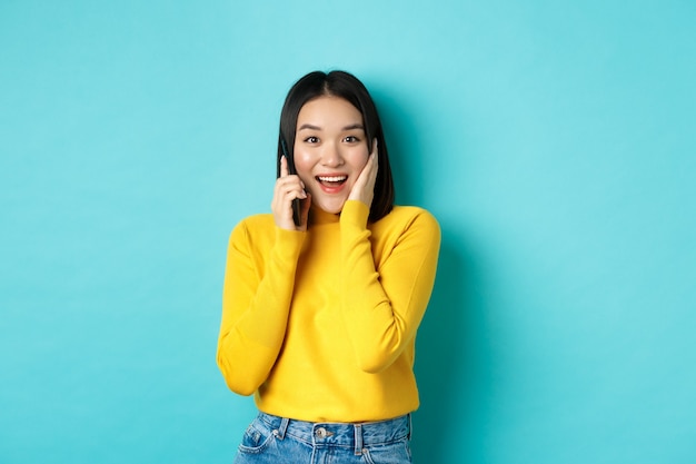 Mulher asiática feliz recebe oferta na ligação, sorrindo enquanto fala no smartphone, em pé sobre um fundo azul.