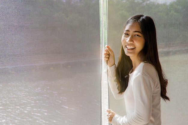 Mulher asiática feliz relaxando com a natureza verde através da janela de vidro em casa