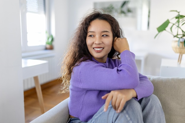 Mulher asiática feliz sentada no sofá em casa e olhando para a câmera Retrato de uma mulher confortável em roupas de inverno relaxando no sofá Retrato de uma mulher bonita sorrindo e relaxando durante o outono