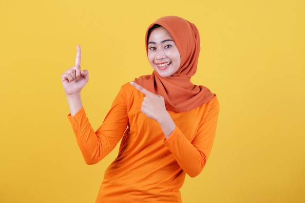 Mulher asiática feliz sorridente com o dedo apontando isolado no fundo da bandeira amarela clara usando hijab