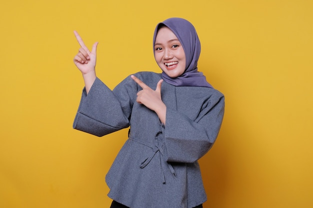 Mulher asiática feliz sorridente usando hijab com o dedo apontando isolado no fundo do banner amarelo claro