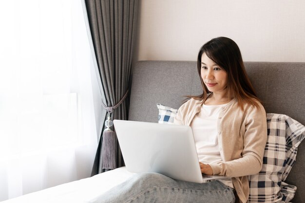 Mulher asiática feliz trabalhando em um laptop sentado na cama. trabalhar a partir do conceito de casa.