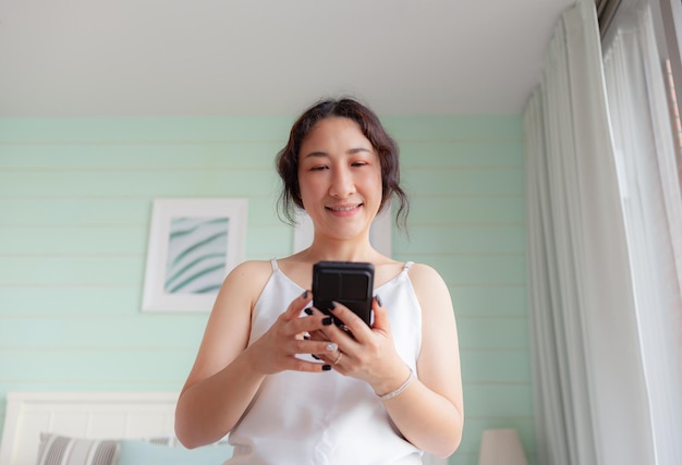 Mulher asiática feliz usando telefone celular em casa Dona de casa asiática verificando mídia social segurando com smartphone relaxa mulher usando aplicativo de celular jogando pedidos de entrega compras videochamada online