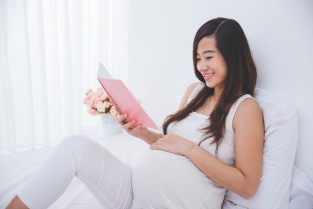 Mulher asiática grávida bonita que lê o livro de nota