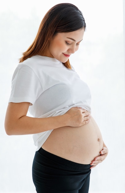 Mulher asiática grávida feliz jovem sorridente vestindo casualmente em pé e tocando sua barriga com amor e carinho, olhando para a câmera para estúdio tiro contra fundo branco.