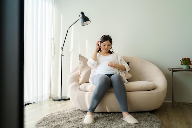 mulher asiática grávida feliz sentada no sofá em casa e usando smartphone para ouvir música