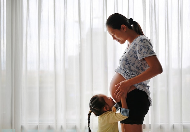 Mulher asiática grávida que está perto da janela em casa com sua filha que beija sua barriga.