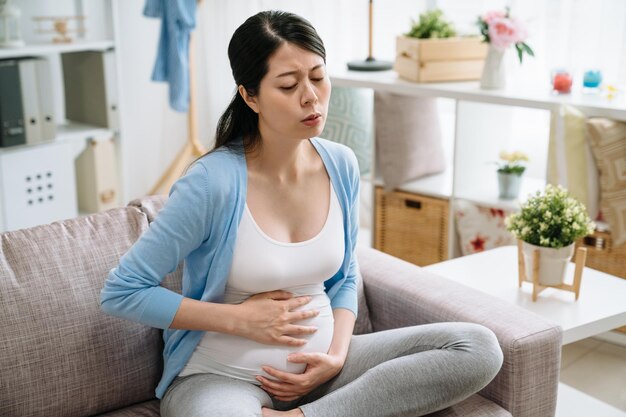 Mulher asiática grávida sofrendo dor de barriga sentada no sofá na sala de estar em casa. jovem tendo bebê futura mãe maternidade paternidade. doença doente fêmea japonesa segurando o estômago doendo.