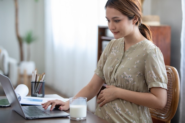 Mulher asiática grávida usando o laptop para trabalhar em casa e bebendo leite