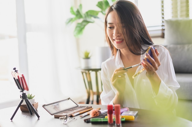 Mulher asiática influenciadora de blogueira de beleza usando streaming ao vivo para smartphone e analisa produtos cosméticos na sala de estar