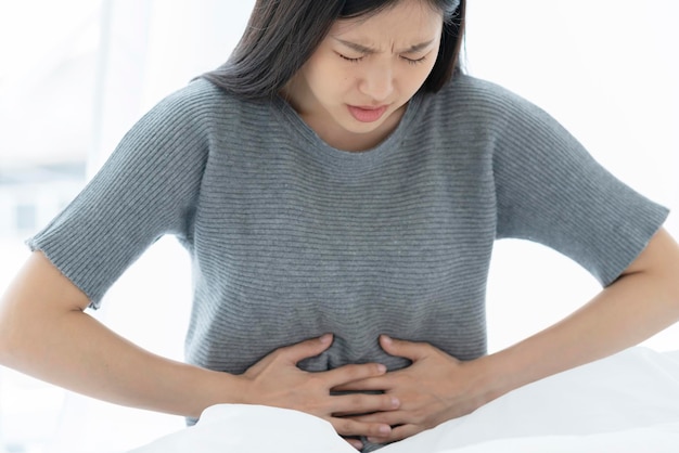 Mulher asiática insalubre toca a mão na barriga, dor de estômago, alergia alimentar ou doente na cama