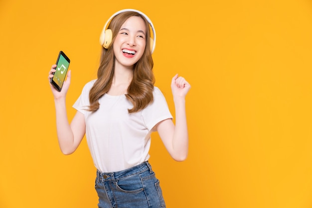 Mulher asiática jovem alegre em fones de ouvido, ouvindo música e desfrute do aplicativo favorito da lista de reprodução no smartphone com dança em fundo laranja.