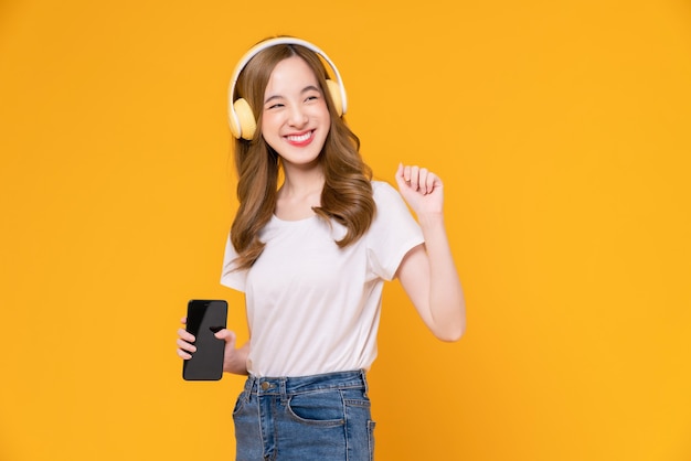 Mulher asiática jovem alegre em fones de ouvido, ouvindo música e desfrute do aplicativo favorito da lista de reprodução no smartphone com dança em fundo laranja.