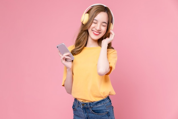 Mulher asiática jovem alegre em fones de ouvido, ouvindo música e desfrute do aplicativo favorito da lista de reprodução no smartphone com dança no fundo rosa.