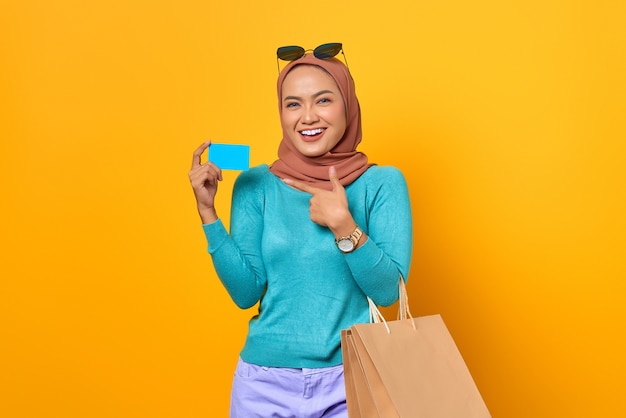 Mulher asiática jovem alegre fazendo compras apontando o dedo para o cartão de crédito em fundo amarelo