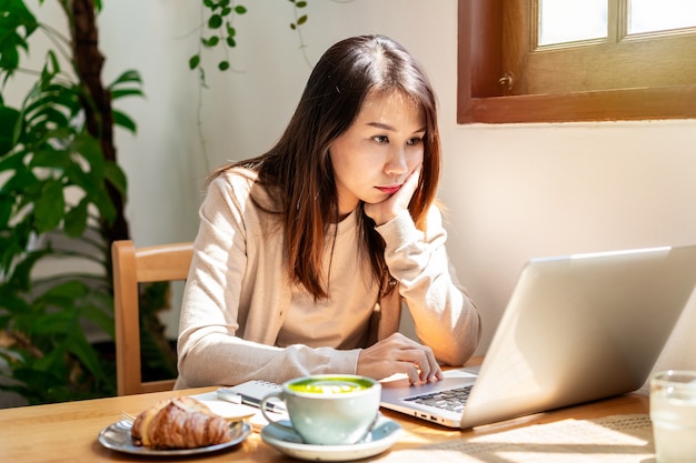 Mulher asiática jovem cansada sentada em um café, trabalhando com uma xícara de café e um croissant